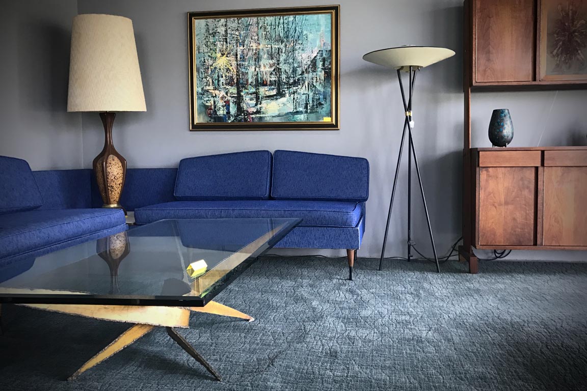 Vintage rich blue upholstered corner platform sofa on a sculptured area rug surrounded with a brutalist steel base and glass coffee table, UFO tripod floor lamp, hardwood wall unit and cork and wood table lamp.