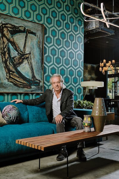 Modern Home 2 Design Showroom owner Mark Davis sits on a couch surrounded by Mid Century Modern furniture, wallpaper, tables, artwork, paintings, sculpture and lighting