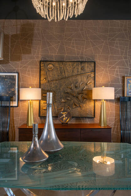 Two polished aluminum metal genie vases sit on a glass top table in front of brass lamps on a wood credenza with fine art paintings on the wall